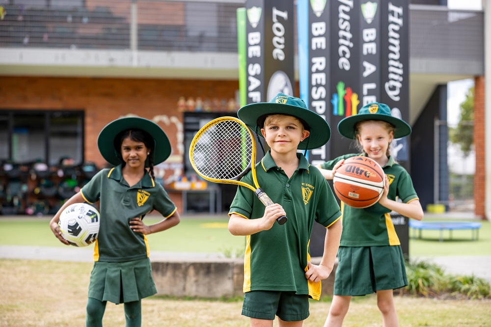 Students in school sports uniforms holding sports equipment outdoors on school grounds.